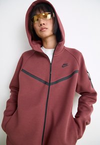 Sweatshirt com fecho éclair da Nike em tecido borgonha com um design de riscas diagonais pretas. Apresenta um capuz grande e bolsos com fecho éclair nas mangas.