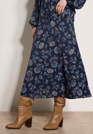 Femme portant une robe midi fleurie bleu marine et des bottines en cuir marron clair à talons mi-mollet, debout sur un sol beige.