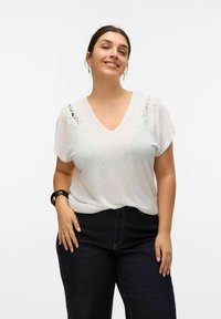 White V-neck top with short sleeves, featuring lace accents on the shoulders, paired with dark blue jeans. Smooth, lightweight fabric.