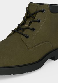 Bottine en daim vert olive avec lacets noirs, œillets métalliques et semelle en caoutchouc noir, présentée en gros plan en angle.