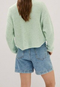 Pull tricoté vert clair à coupe ample avec motif texturé, associé à un short en jean bleu taille haute avec poches arrière.