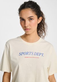 Cremefarvet t-shirt med "SPORTS DEPT." i blå og "hummel PERFORMANCE" i rød. Korte ærmer, rund hals, blød tekstur.