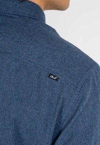 Blaues Flanellhemd mit strukturiertem Stoff, Button-Down-Kragen und kleinem schwarzen Markenschild auf der oberen Rückseite.