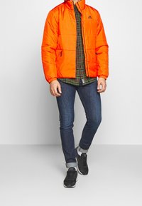 Orange puffjacka med hög krage, med dragkedjefickor och ett broderat logotyp, bärd över en rutig skjorta, parat med mörka jeans och sneakers.