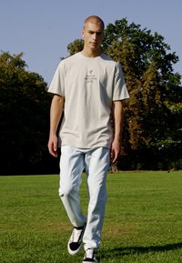 Beige cotton t-shirt with front text, light blue jeans, and black and white sneakers. Walking on grass with trees in the background.