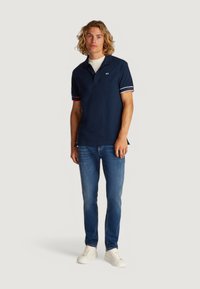Polo navy con cuello clásico, que presenta acentos a rayas rojas y blancas en las mangas y un detalle de logo. Llevado con vaqueros azules y zapatos blancos.