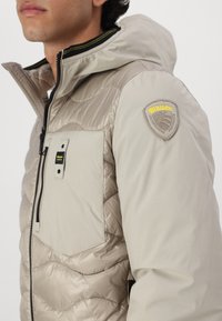 Giacca trapuntata beige chiaro con cappuccio, dotata di tasca sul petto e di una patch con logo sulla spalla. Texture liscia, cerniera nera a contrasto.