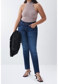 Mujer con top sin mangas color malva, vaqueros de talle alto azul y tacones negros con tiras, sosteniendo una chaqueta negra sobre un fondo liso.