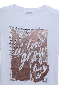 T-shirt bianca con una grafica metallica rosa oro e la scritta "Fai fiorire l'amore" accompagnata da un cuore. Testo in stile calligrafico nero lo circonda.