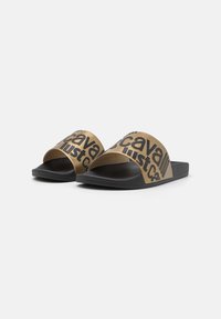 I sandali slide dorati presentano un'ampia fascia con lettering nero e una suola in gomma nera testurizzata. Design durevole e minimalista per un look informale.