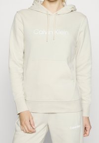 Sweat à capuche en coton beige avec une poche avant, une capuche à cordon et le logo blanc "Calvin Klein". Présente une texture lisse et des poignets côtelés.