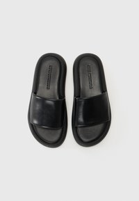 Sandales mules en faux cuir noir avec une large bride, un bout rond et une semelle rembourrée, présentant un logo embossé sur le côté.
