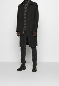 Hombre vestido con un abrigo negro largo, jeans negros, zapatillas negras y un Suéter de cuello alto a rayas negras y grises, de pie frente a una pared blanca lisa.