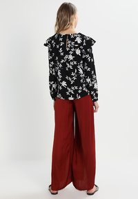 Blusa nera floreale con motivi bianchi a foglia e maniche a volant, abbinata a pantaloni larghi rossi. La vista posteriore mostra un'apertura a chiave.