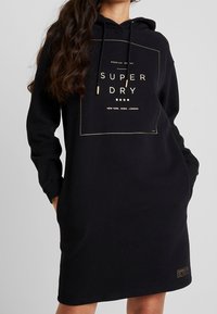 Kvinna som bär en svart huvtröja med "Super Dry"-logotyp och stadnamn tryckta i guld, händerna i fickorna, långt vågigt hår över axeln.
