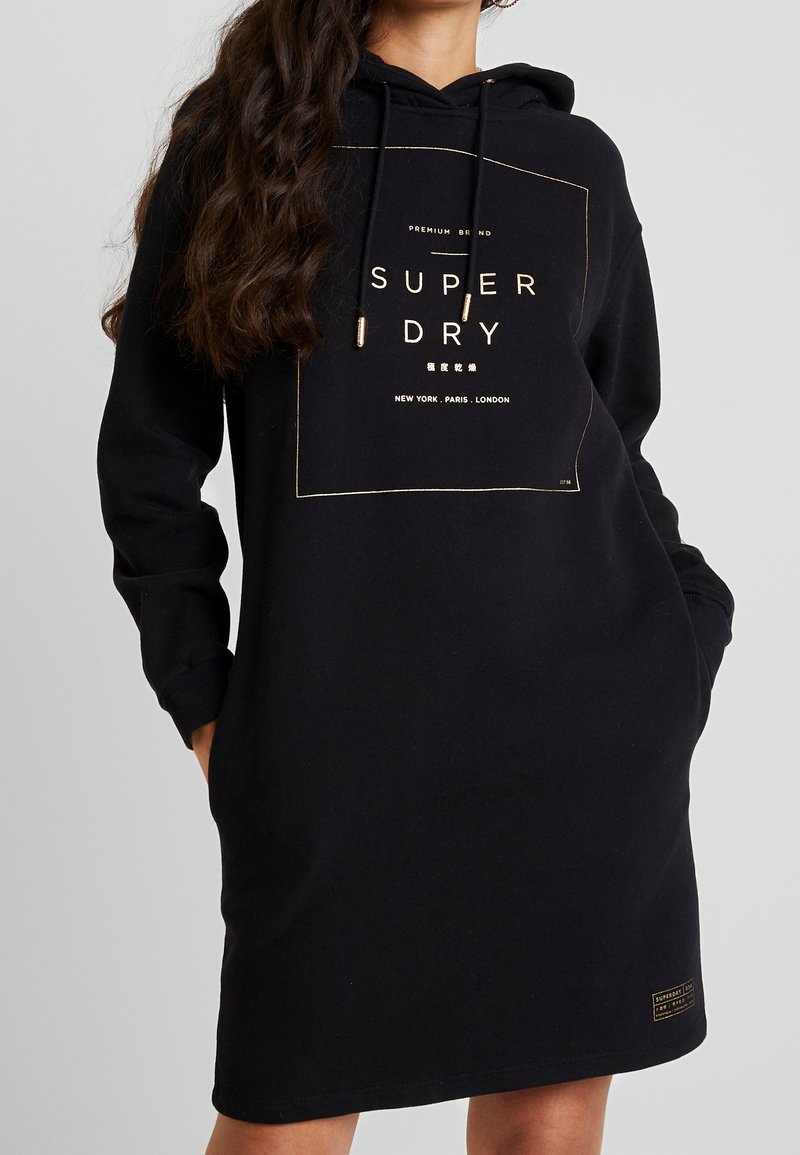 Kvinna som bär en svart huvtröja med "Super Dry"-logotyp och stadnamn tryckta i guld, händerna i fickorna, långt vågigt hår över axeln.
