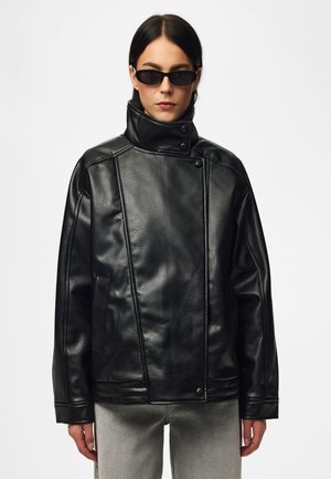 Femme portant une veste en cuir noire à col montant et des lunettes de soleil noires sur un fond clair uni.