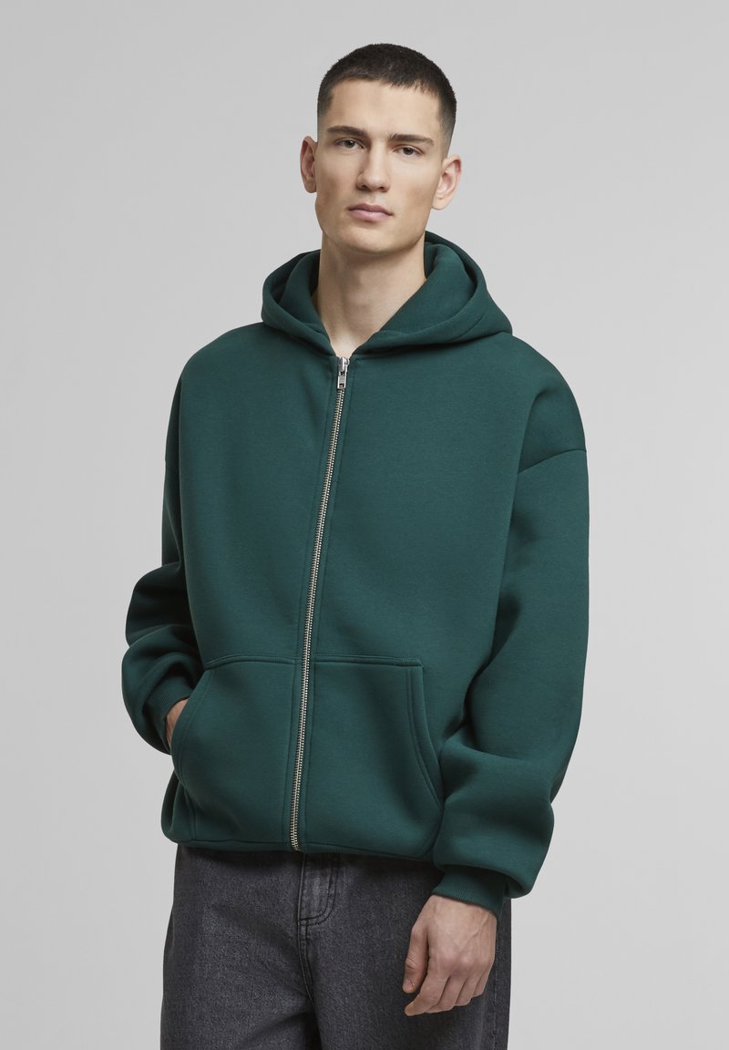 Mørkegrøn zip-up hoodie med foret hætte, frontlommer og ribbede manchetter. Lavet af blødt, tykt stof med en afslappet pasform.