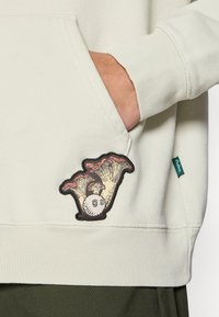 Main d'une personne dans la poche d'un sweat crème avec un patch en forme de champignon et de balle de golf sur la poche, petite étiquette en tissu vert visible sur la couture latérale.