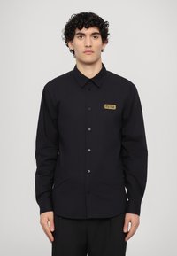 Black long-sleeve shirt featuring a button-up design, a fold-down collar, and a gold logo patch on the left chest. Smooth cotton fabric.