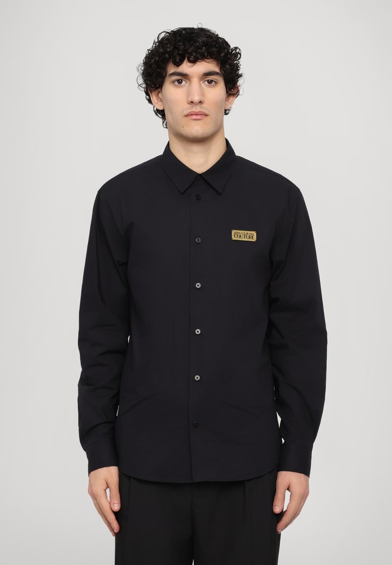 Black long-sleeve shirt featuring a button-up design, a fold-down collar, and a gold logo patch on the left chest. Smooth cotton fabric.