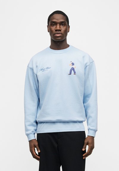 Νέος άνδρας που φοράει ανοιχτό μπλε φούτερ με κεντημένη φιγούρα περιπάτου που κρατά ψωμί και σκούρα παντελόνια, στέκεται μπροστά σε απλό φόντο.