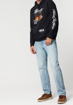 Personne portant un sweat à capuche graphique noir avec du texte blanc, un jean bleu clair et des baskets en daim marron, debout devant un fond blanc uni.
