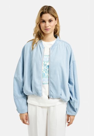 Hellblaues Denim-Bomberjackett mit einem Reißverschluss vorne, herunterhängenden Schultern und elastischen Bündchen. Getragen über einem weißen T-Shirt mit floralem Design.