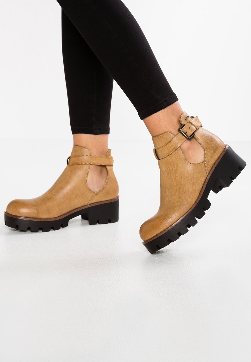 Bottes en cuir beige montant à la cheville avec semelles noires épaisses et sangles à boucle, portées avec des leggings noirs, photographiées en train de marcher sur un sol blanc.