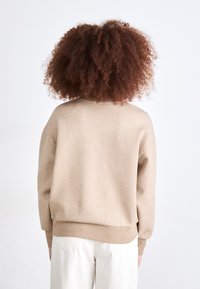 Beige genser med avslappet passform, ribbede mansjetter og kant, teksturert stoff og rund hals, vist fra bakgrunn mot en lys bakgrunn.