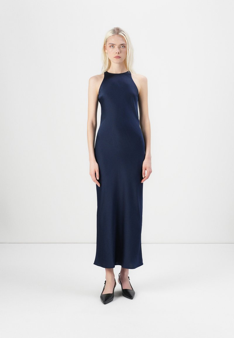 Mørkeblå halterneck maxi kjole med et glat, strømlinet stof. Flade sko med spidse tæer supplerer det minimalistiske design.