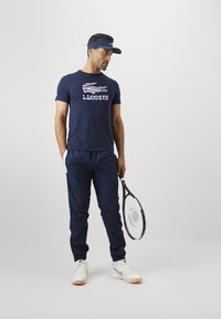 Homem de t-shirt náutico da Lacoste, calças náuticas, ténis brancos e viseira, segurando uma raquete de ténis, em pé contra um fundo claro e liso.