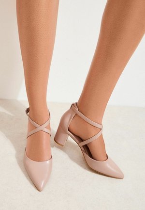 Femeie purtând pantofi cu toc gros de culoare nude, cu vârf ascuțit și barete încrucișate la gleznă, stând pe un podea deschisă la culoare.