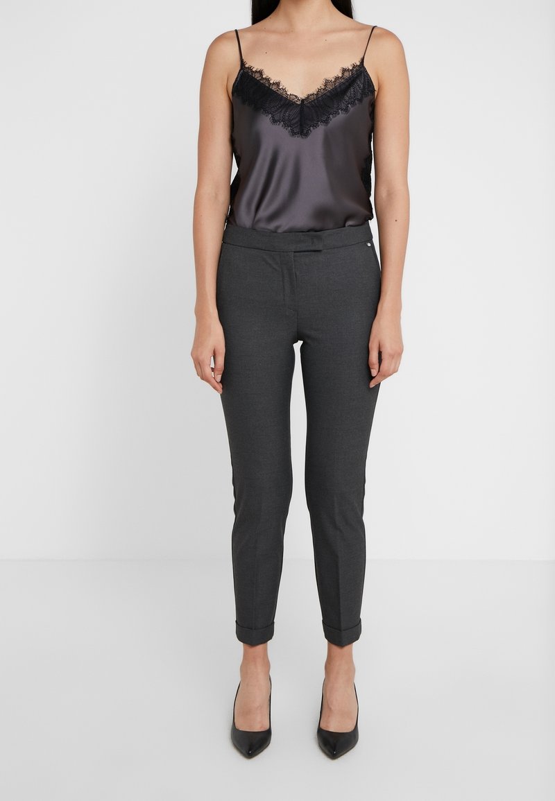 Femme portant un caraco en satin noir orné de dentelle, un pantalon slim gris foncé avec ourlets repliés, et des escarpins noirs à bout pointu, debout devant un fond blanc.