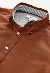 Camicia in cotone marrone con colletto standard, dotata di bottoni bianchi e dettagli a righe all'interno del colletto. Piccolo logo ricamato sul petto.