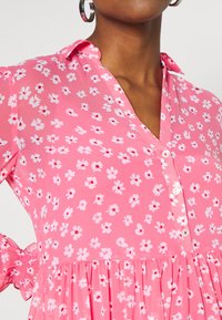 Abito rosa con motivo floreale, colletto a V con bottoni, vita arricciata e maniche voluminose, realizzato in un tessuto leggero con piccoli fiori bianchi.