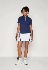 Mulher de pé, vestindo uma polo azul-marinho e uma saia de golfe branca, segurando um taco de golfe, com ténis brancos e meias, contra um fundo simples.