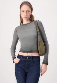 Sivi dugi rukav crops top s izvezenim tekstom, uparen s tamnoplavim trapericama. Torba na rame u maslinastozelenoj antilop koži prikazana s jedne strane.