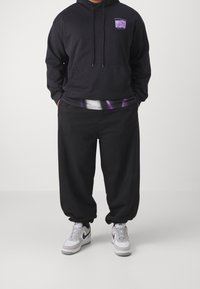 Person trägt einen schwarzen Kapuzenpullover und lockere schwarze Jogginghosen mit weißen und grauen Nike-Sneakern, die Hände in den Taschen, vor einem schlichten Hintergrund.