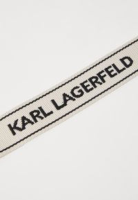 Hvit vevet bånd med "KARL LAGERFELD" brodert med fete sorte bokstaver; har en teksturert overflate og sorte kantkanter.