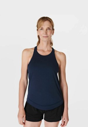 Vrouw met licht haar, die een marineblauw mouwloos topje en zwarte korte broek draagt, staand tegen een effen witte achtergrond.