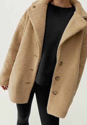 Femme portant un manteau teddy beige à grand col et boutons, par-dessus un sweat-shirt et un pantalon noirs, debout devant un fond uni.