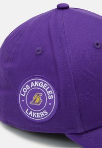 Purple baseball cap with Los Angeles Lakers logo patch, featuring flat brim and top button. Made from textured fabric with stitched details.