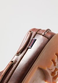 Braunes Leder-Schuhwerk mit Metallösen, ausgestattet mit einem gestickten Design, einer strukturierten Gummisohle und einem roten und blauen Etikett an der Seite.