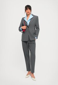 Woman wearing a gray pantsuit with a light blue shirt, holding a small red purse, and wearing white pointed-toe slingback heels.