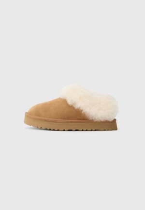 Pantofole in suede marrone a slip-on con un spesso risvolto in shearling beige e suola in gomma, su sfondo bianco uniforme.