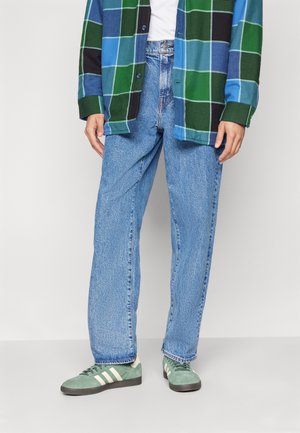 Persona che indossa una camicia a quadri blu-verde, jeans dritti azzurro chiaro e scarpe da ginnastica Adidas verdi con strisce bianche, in piedi su un pavimento semplice.