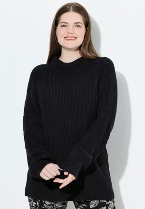 Suéter negro de punto con cuello alto y mangas de punto texturizado en relieve. Combinado con pantalones oscuros de patrón que presentan motivos de hojas.