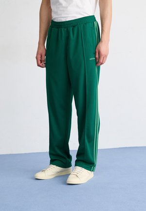 Person trägt dunkelgrüne Adidas-Trainingshose mit hellgrünen Seitenstreifen und weißen Sneakern, steht auf einem hellblauen Boden.