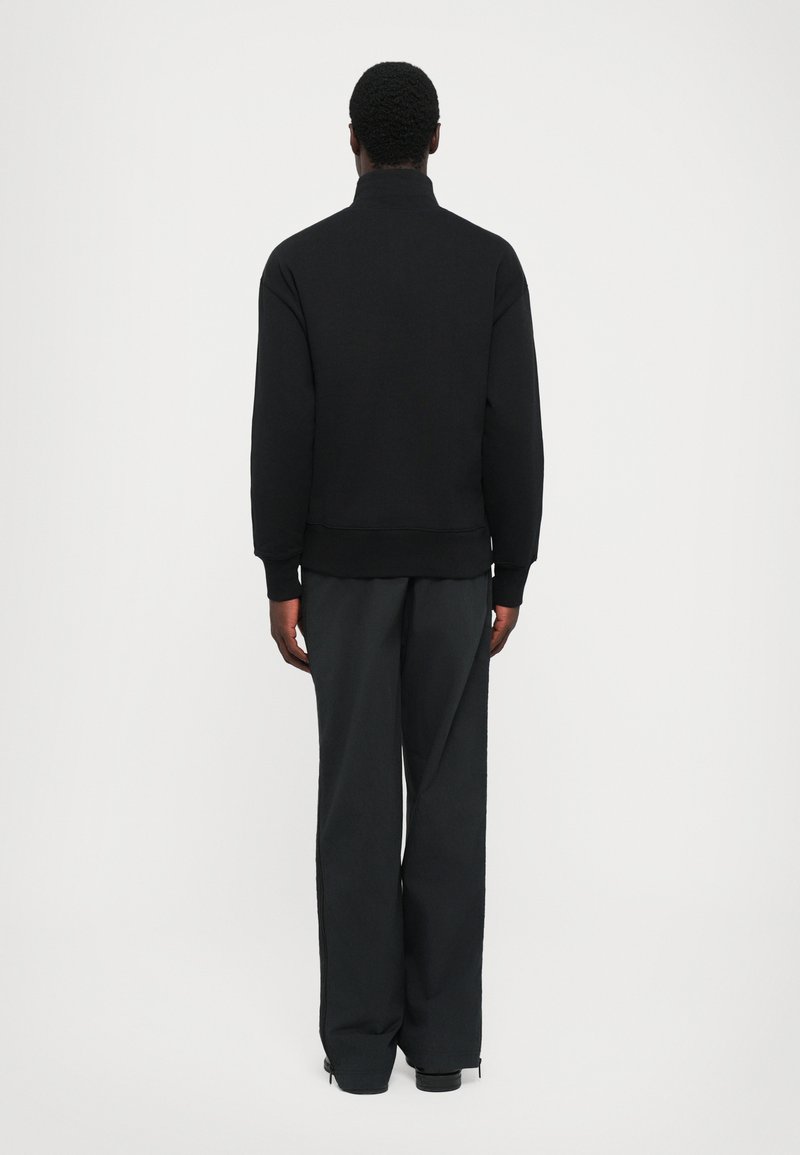 Man die met zijn gezicht weg staat, gekleed in een zwarte hooggesloten trui en losse zwarte broeken, tegen een eenvoudige witte achtergrond.